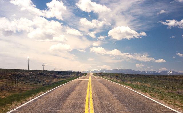 Looking down a long empty road in the middle of nowhere with double yellow lines. Leading to distant snow capped mountains.